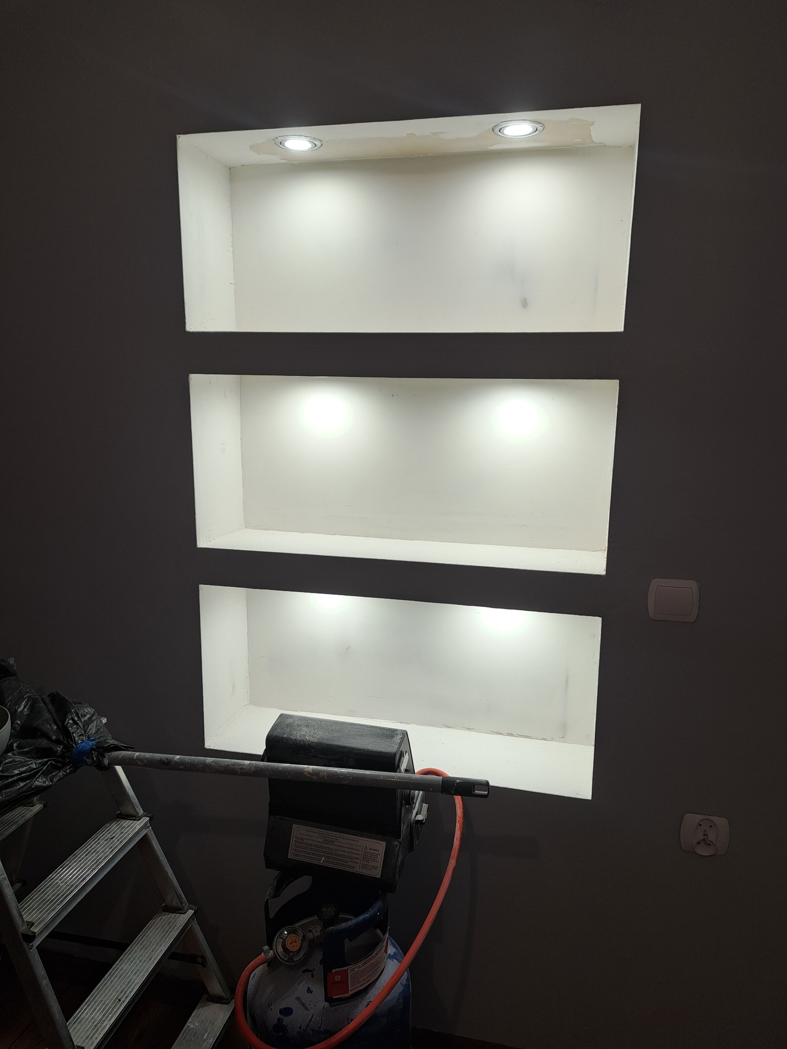 Ściana w odcieniach szarości z trzema wnękami oświetlonymi punktowo. Widoczny sprzęt malarski i drabina, sugerujące etap prac wykończeniowych.