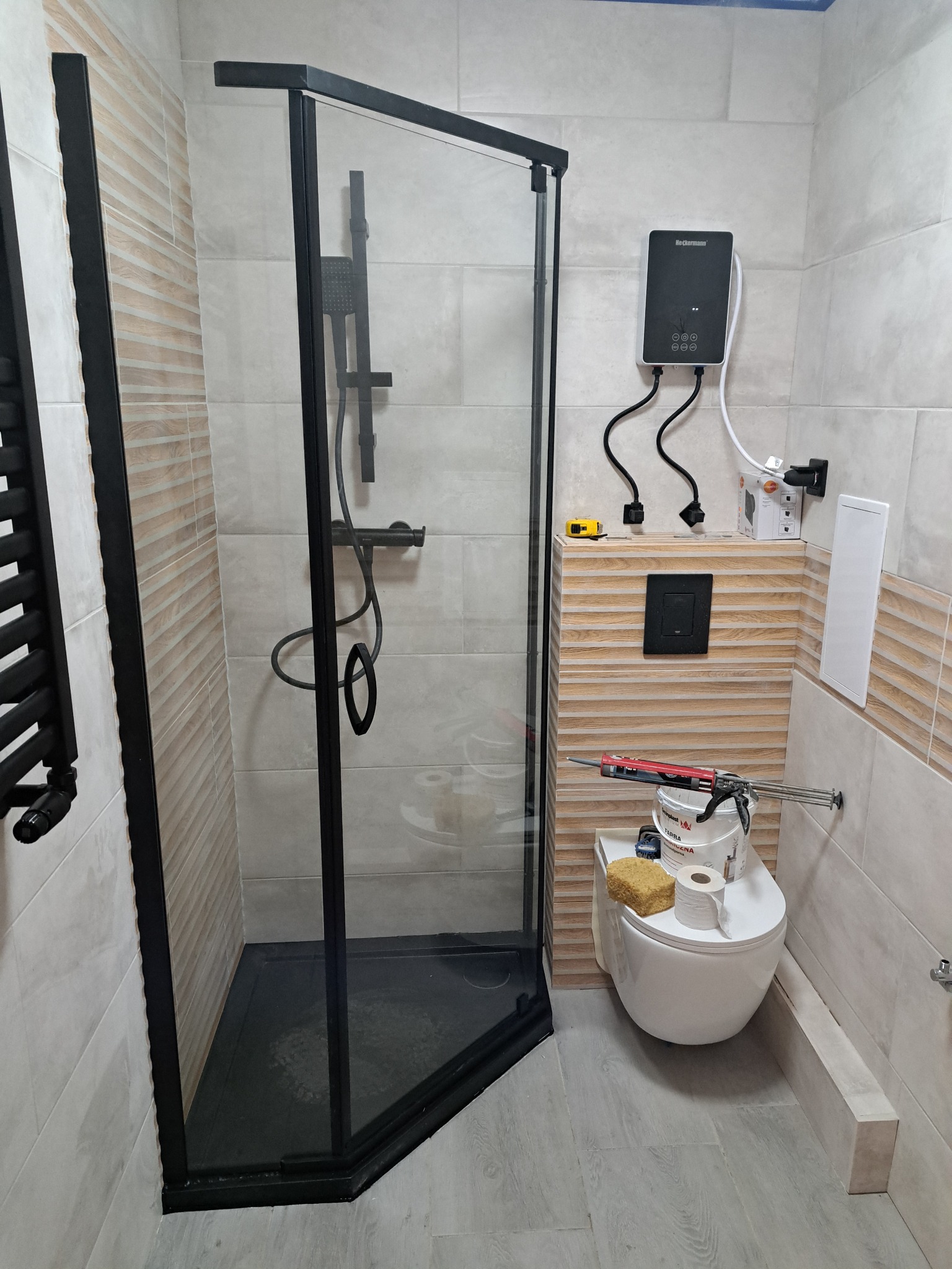 Nowoczesna kabina prysznicowa z czarnymi profilami w wykończonej łazience. Widoczne narzędzia i materiały budowlane sugerują etap prac wykończeniowych.