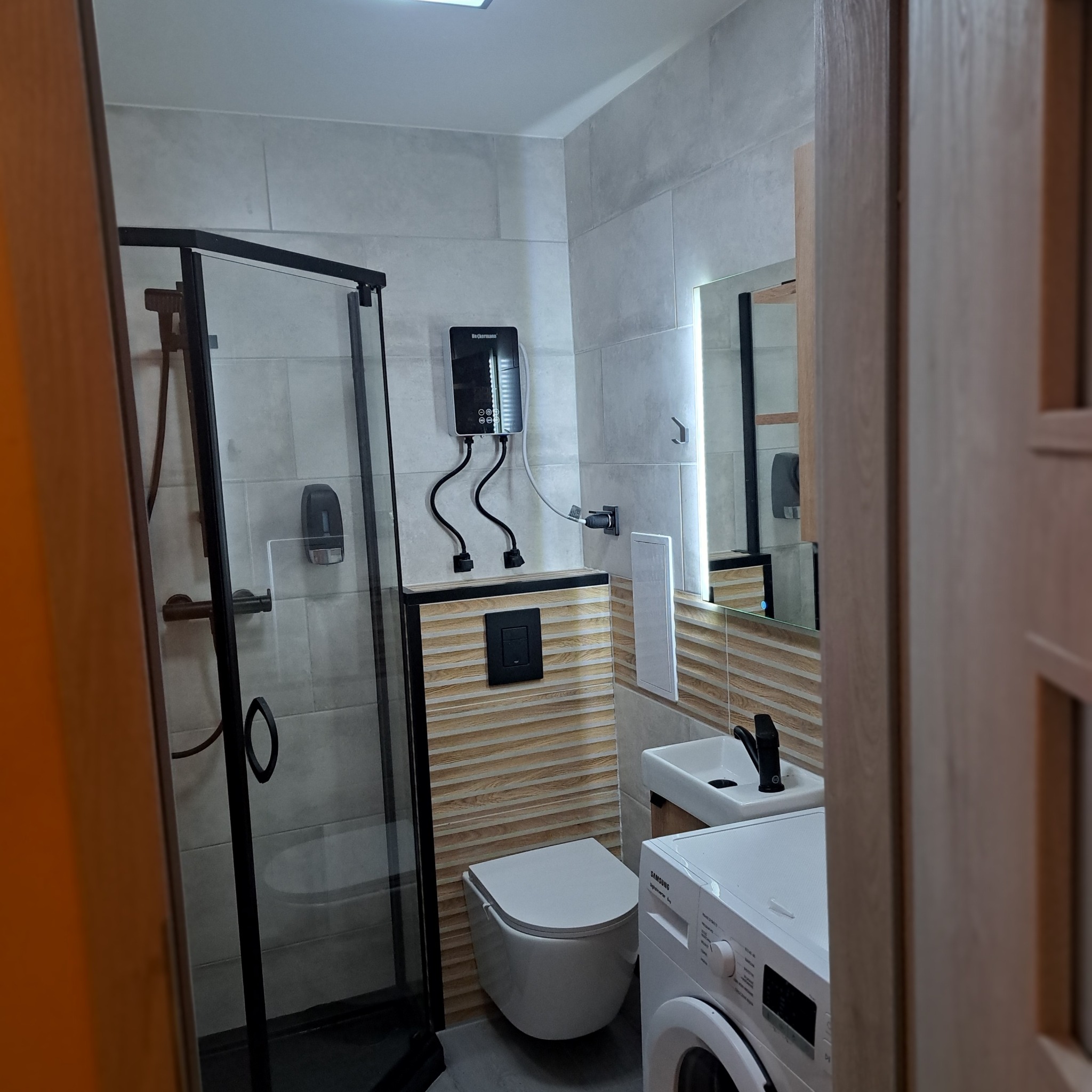 Nowoczesna łazienka z kabiną prysznicową typu walk-in, podwieszaną toaletą i pralką Samsung. Ściany wyłożone szarymi płytkami z dekoracyjnym panelem.