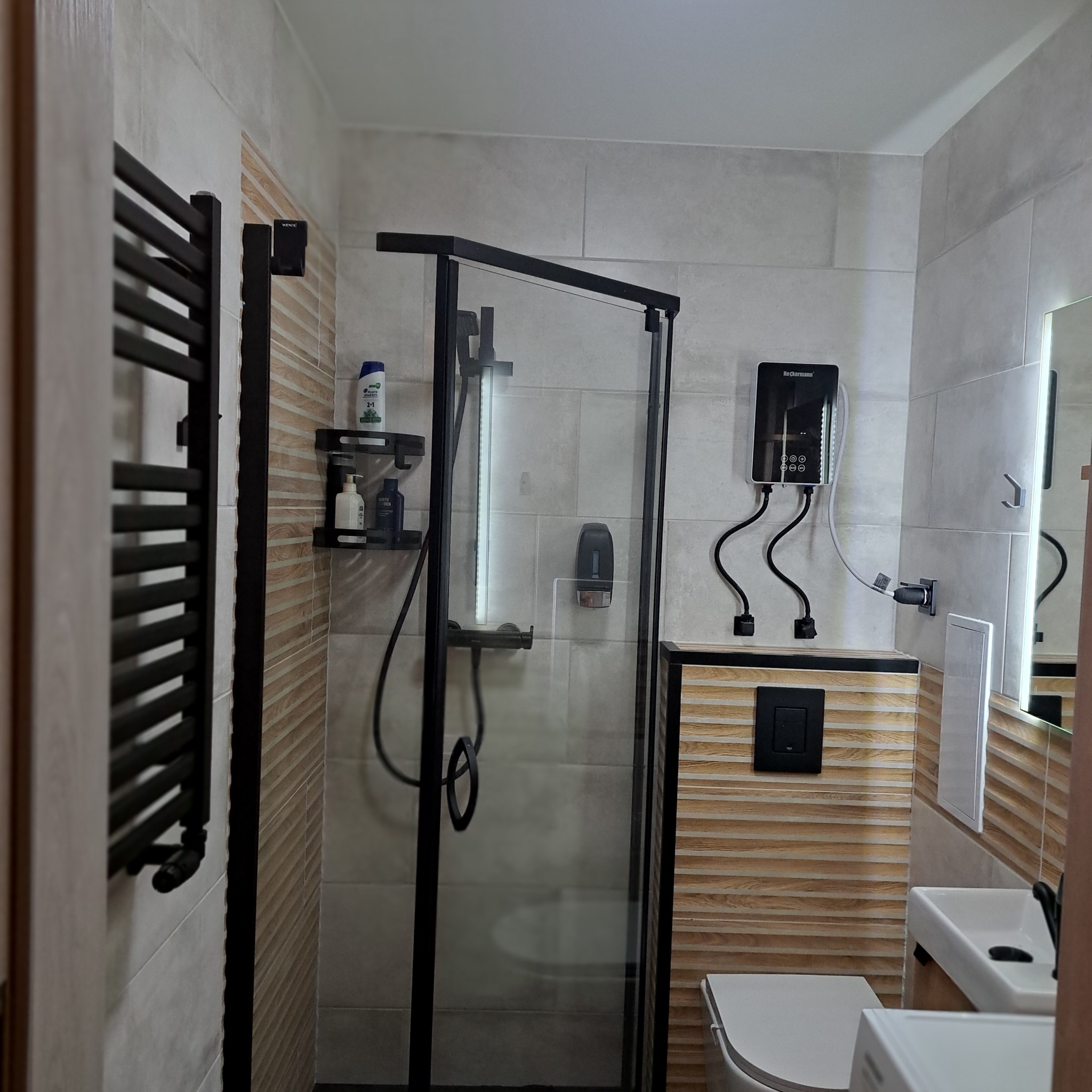 Nowoczesna łazienka po remoncie: kabina prysznicowa z czarną ramą, grzejnik drabinkowy, beżowe płytki imitujące drewno i szare płytki. Widoczny podgrzewacz wody.