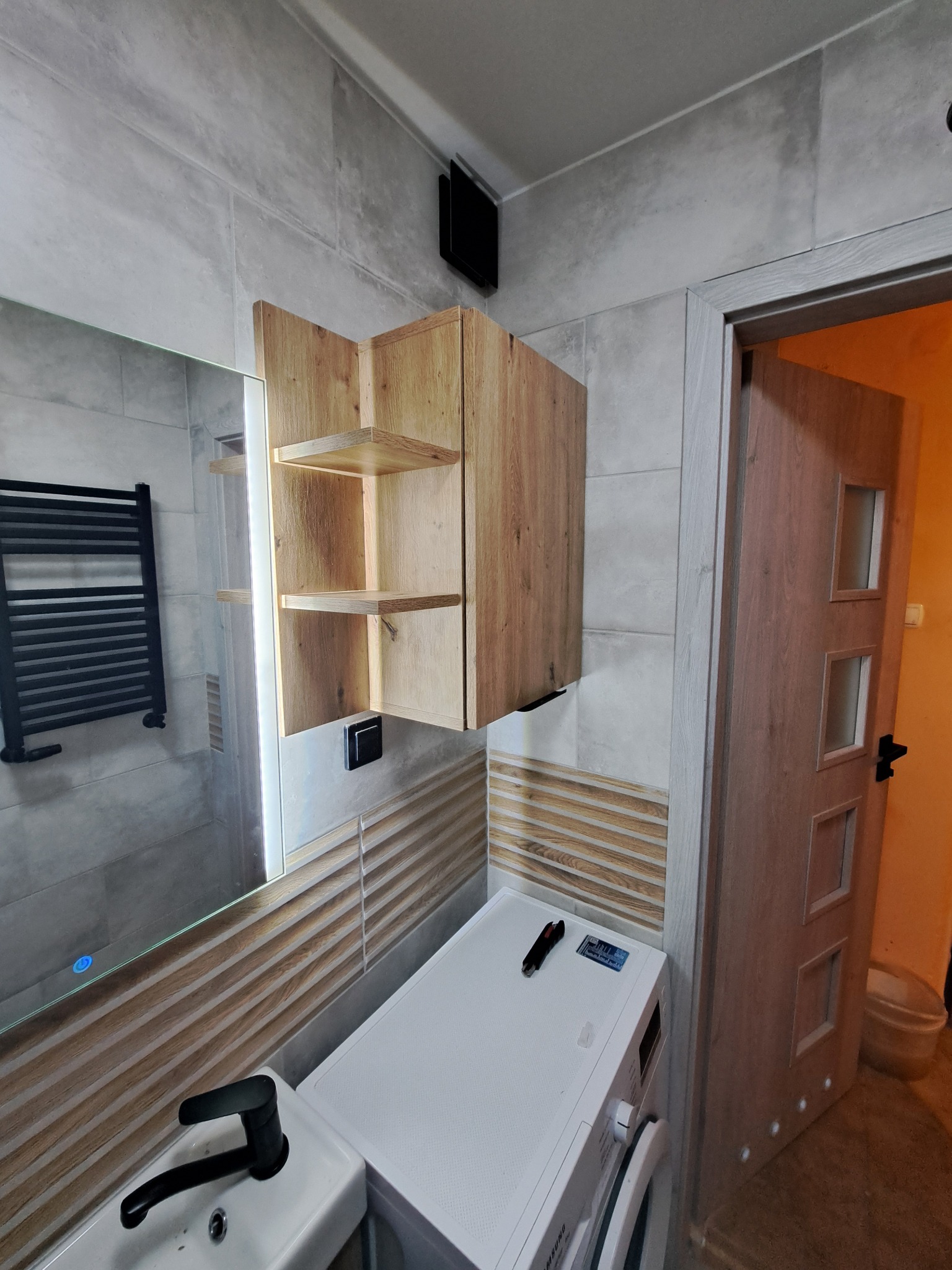 Nowoczesna łazienka z drewnianą szafką nad pralką, lustrem z podświetleniem i grzejnikiem drabinkowym. Uwagę zwracają płytki imitujące drewno na ścianie.