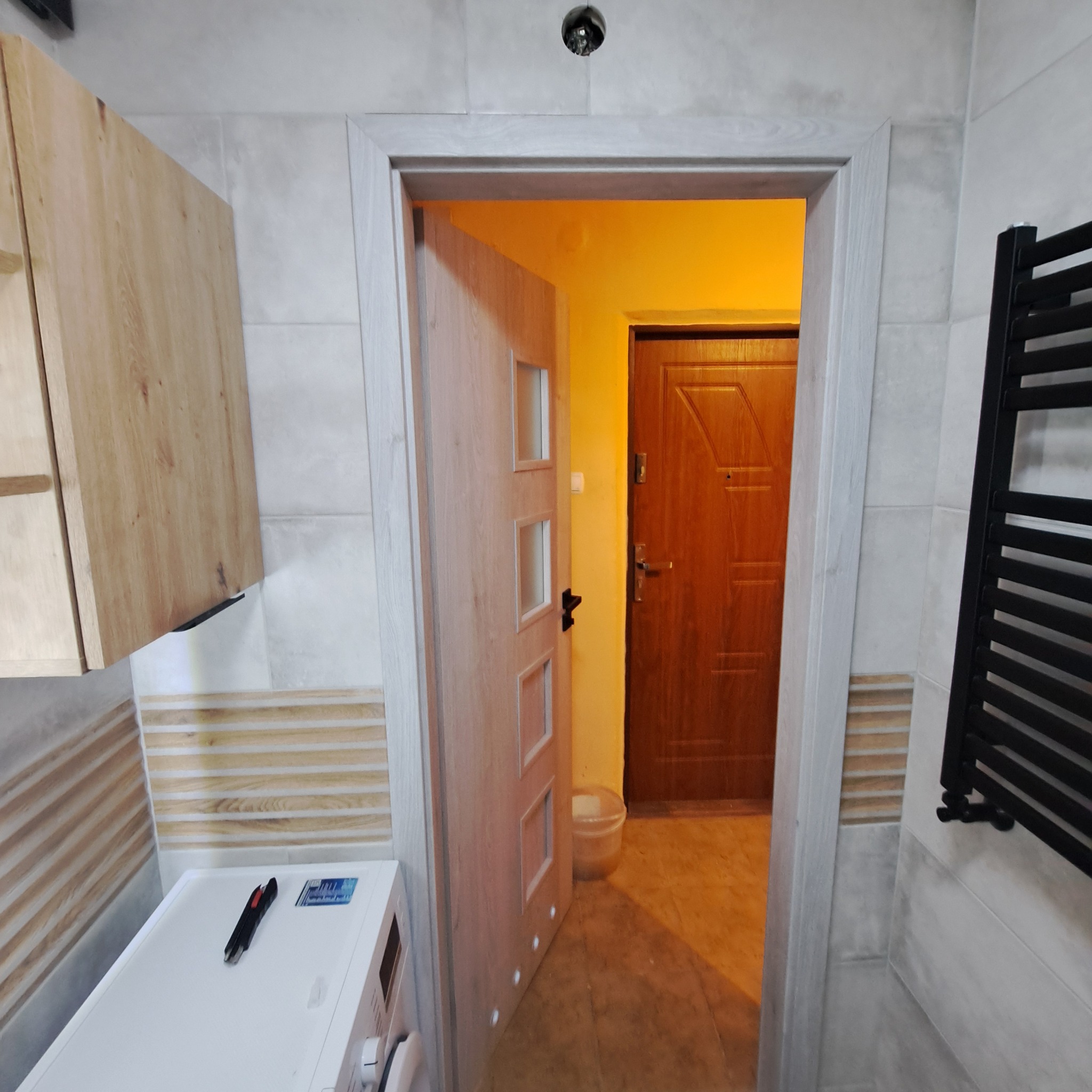 Wykończona łazienka z drewnianą szafką, pralką i grzejnikiem drabinkowym. Widoczne płytki imitujące drewno i otwarta drewniana rama drzwi z widokiem na kolejne pomieszczenie.