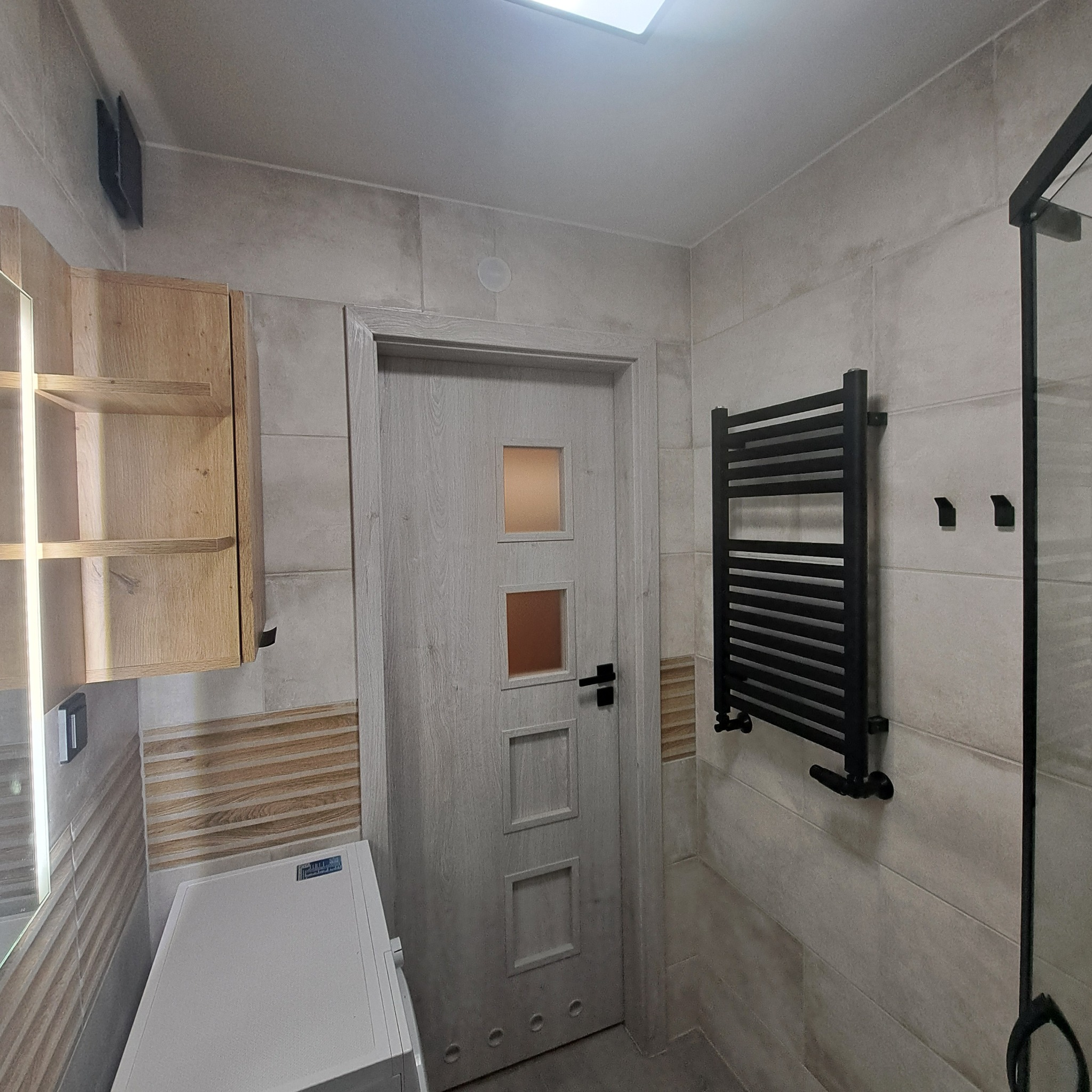 Wykończona łazienka w beżowych płytkach z drewnianymi akcentami. Widoczna pralka, grzejnik drabinkowy i drzwi z przeszkleniami. Oświetlenie sufitowe.