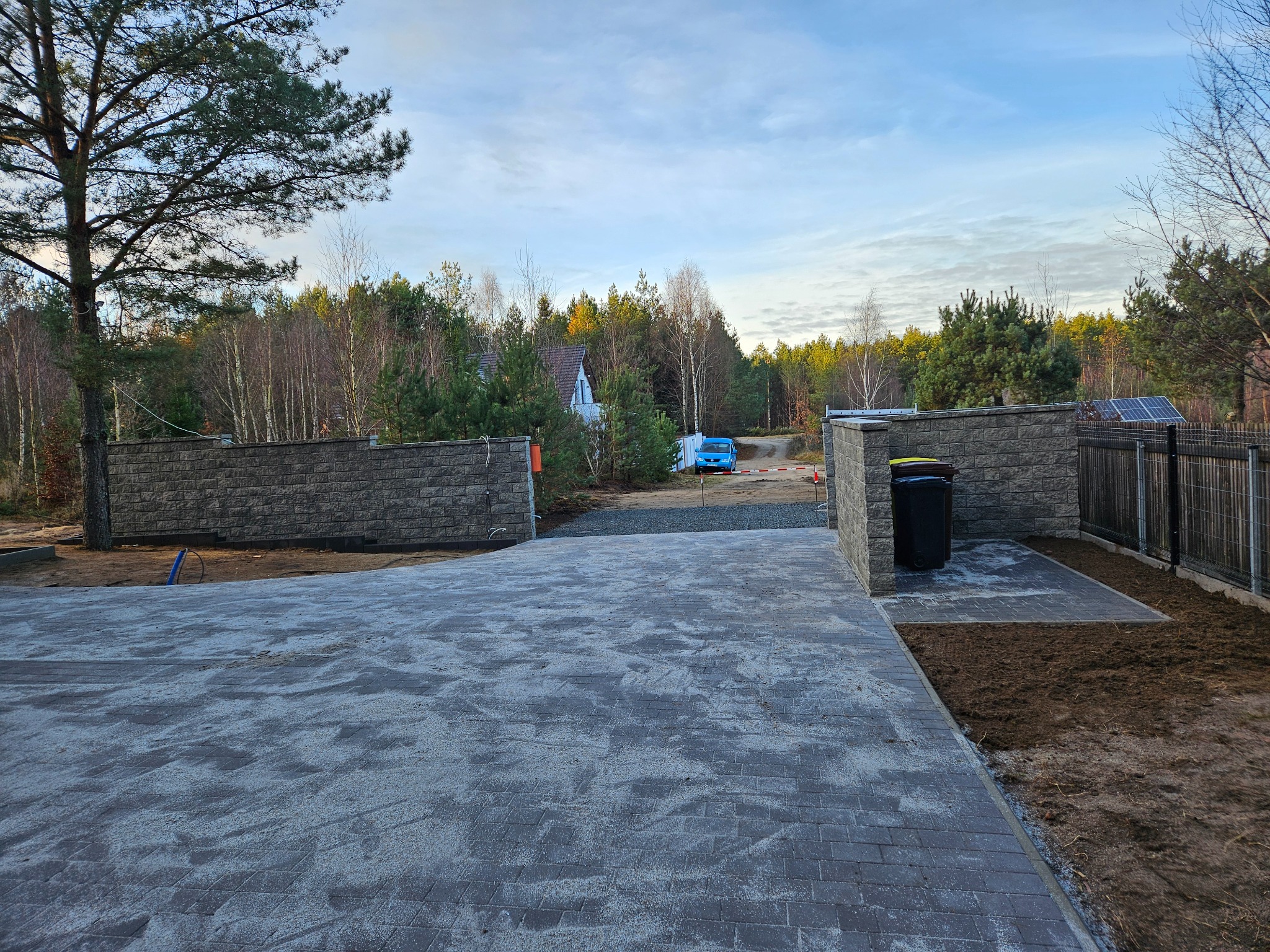 Ułożona kostka brukowa na podjeździe, z widocznym fragmentem ogrodzenia z kamienia łupanego i siatki. W tle las i błękitny samochód.
