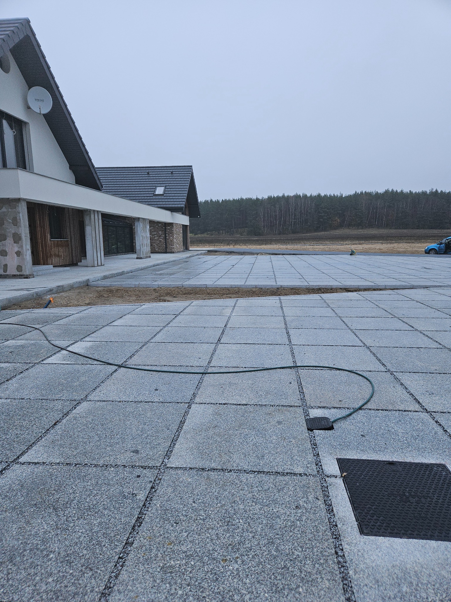 Ułożona szara kostka brukowa na posesji domu jednorodzinnego. Widoczny fragment budynku, tło z drzewami i niebieski samochód w oddali. Kostka z widocznymi fugami.