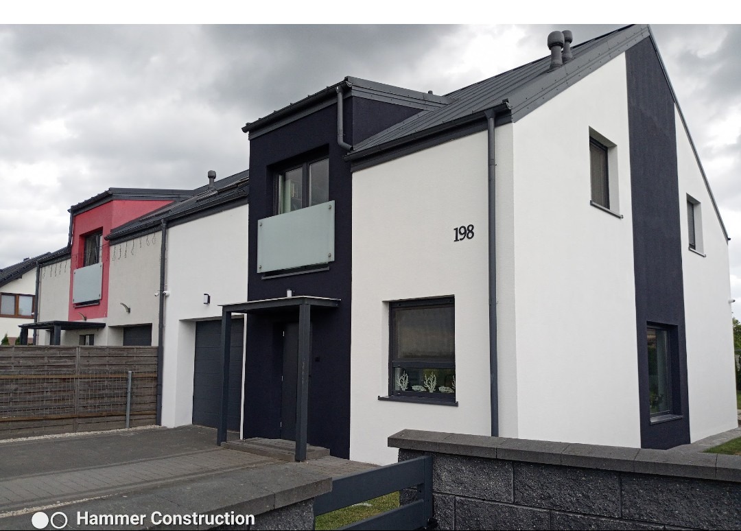 Nowoczesny dom jednorodzinny z elewacją w kolorach bieli i grafitu, numer 198 na ścianie. Widok z zewnątrz, w tle inne domy w zabudowie szeregowej.