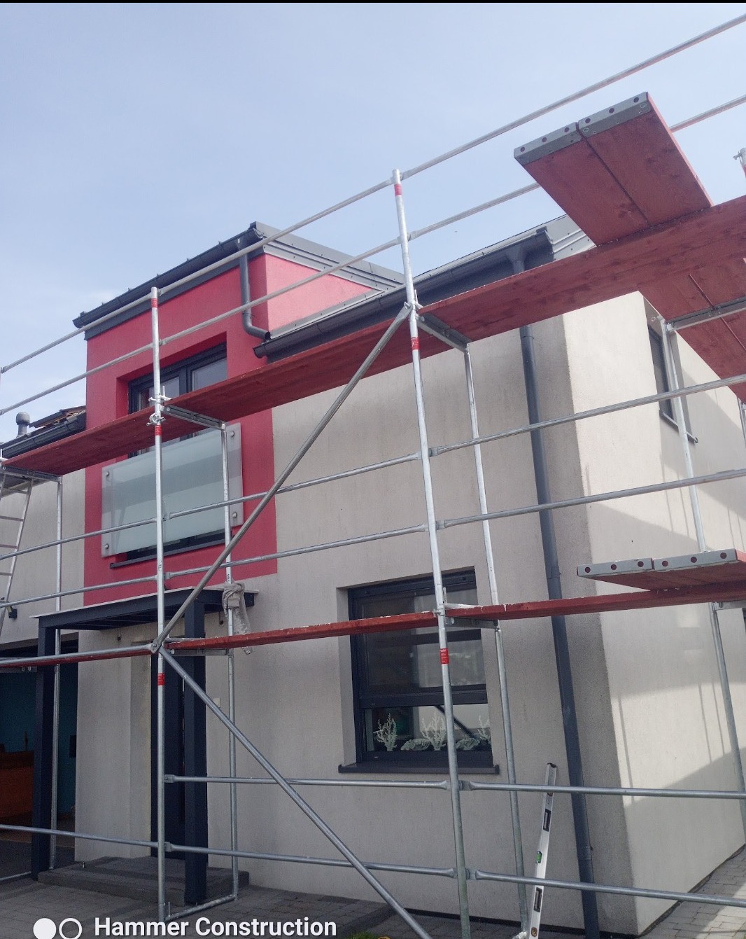 Dwukondygnacyjny dom w trakcie prac elewacyjnych, z widocznymi rusztowaniami i fragmentami pomalowanej fasady na czerwono. Ujęcie z dołu, podkreślające wysokość budynku.