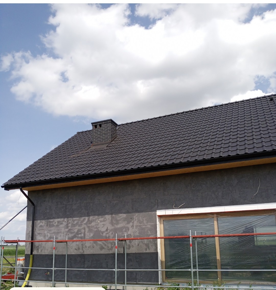 Dom w trakcie prac elewacyjnych: widoczne rusztowania, okno zabezpieczone folią i fragment ocieplonej ściany z szarymi panelami. Dach pokryty ciemną dachówką.