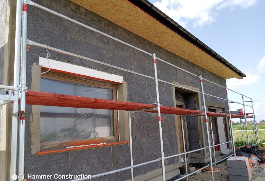Budynek w trakcie prac elewacyjnych, widoczne rusztowania, okna zabezpieczone folią, ściany z szarego bloczka, fragment dachu z deskowaniem.