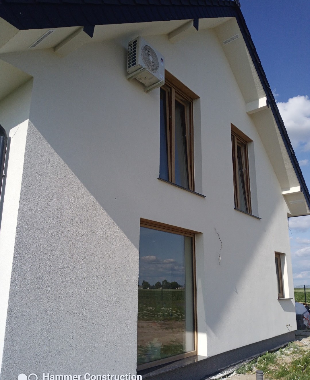 Biała elewacja domu jednorodzinnego z drewnianymi oknami i klimatyzatorem. Widoczna faktura tynku i fragment dachu. W oknach odbija się krajobraz.