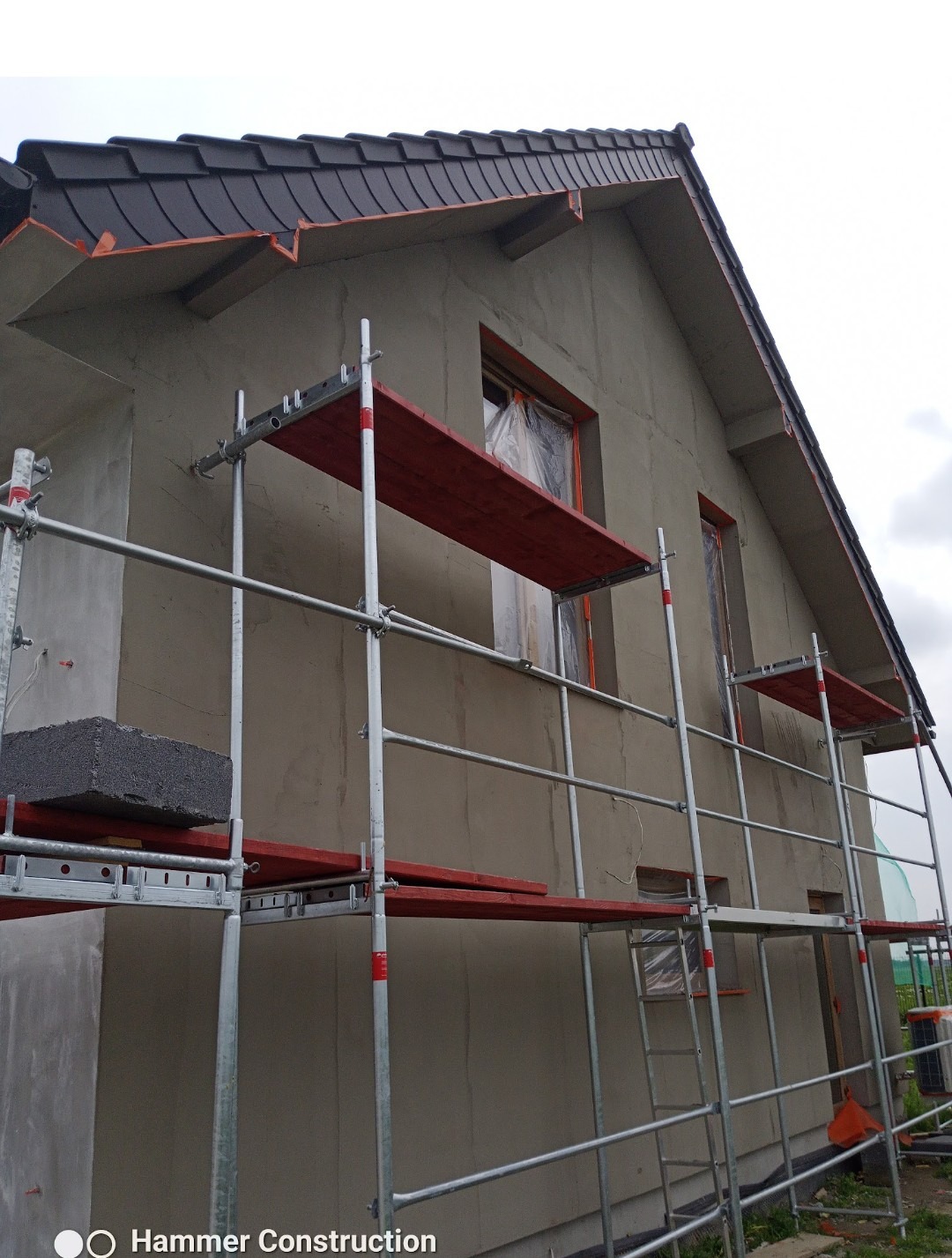 Dom w trakcie prac elewacyjnych: szare ściany, rusztowania z czerwonymi elementami, okna zabezpieczone folią. Widoczny fragment dachu z ciemną dachówką.