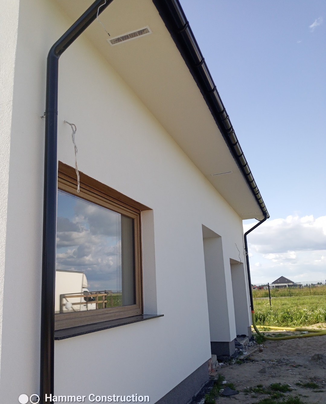 Boczny widok otynkowanego domu jednorodzinnego z brązowymi oknami, czarną rynną i widocznymi elementami wykończeniowymi elewacji. W tle zielona trawa i błękitne niebo.