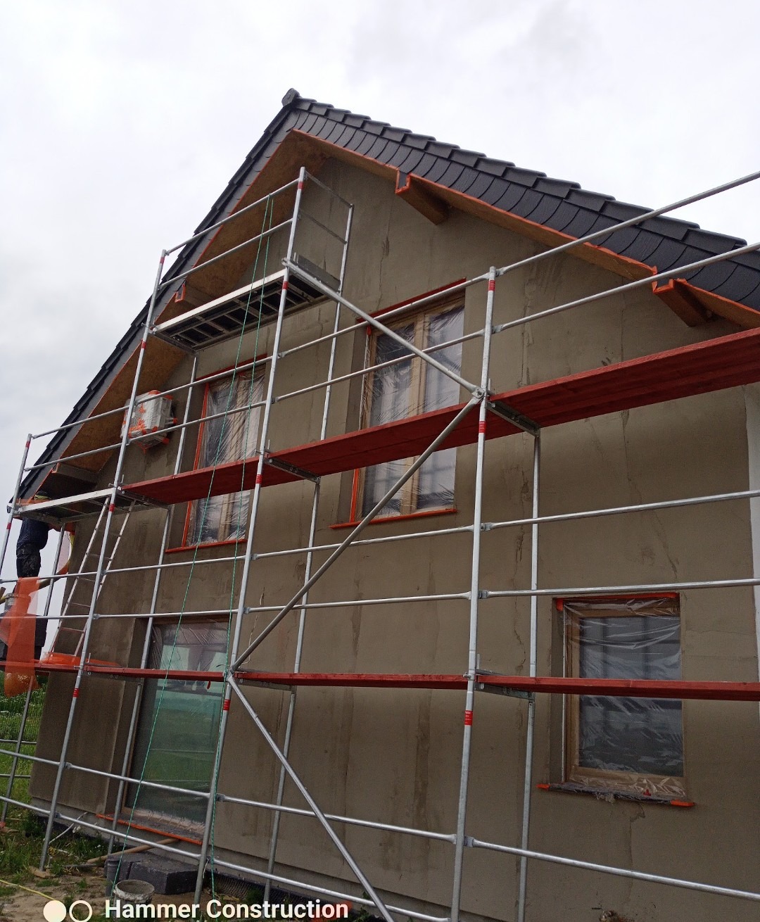 Dom w trakcie nakładania tynku zewnętrznego, widoczne rusztowania i okna zabezpieczone folią. Ściana w szarym kolorze, dach pokryty ciemną dachówką.