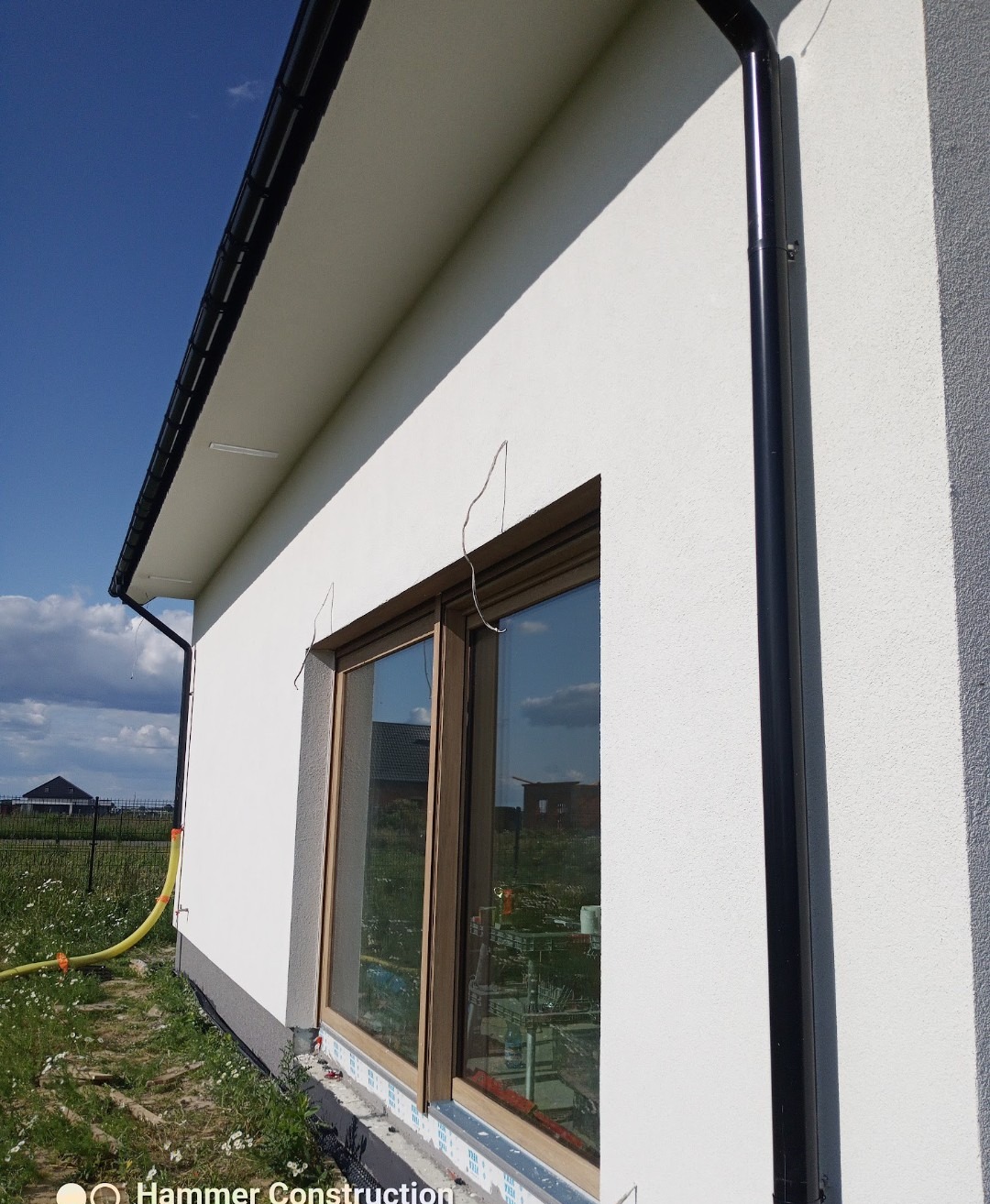 Biała elewacja domu jednorodzinnego z drewnianą stolarką okienną i czarną rynną spustową. Widoczny fragment trawnika i niebo z chmurami.