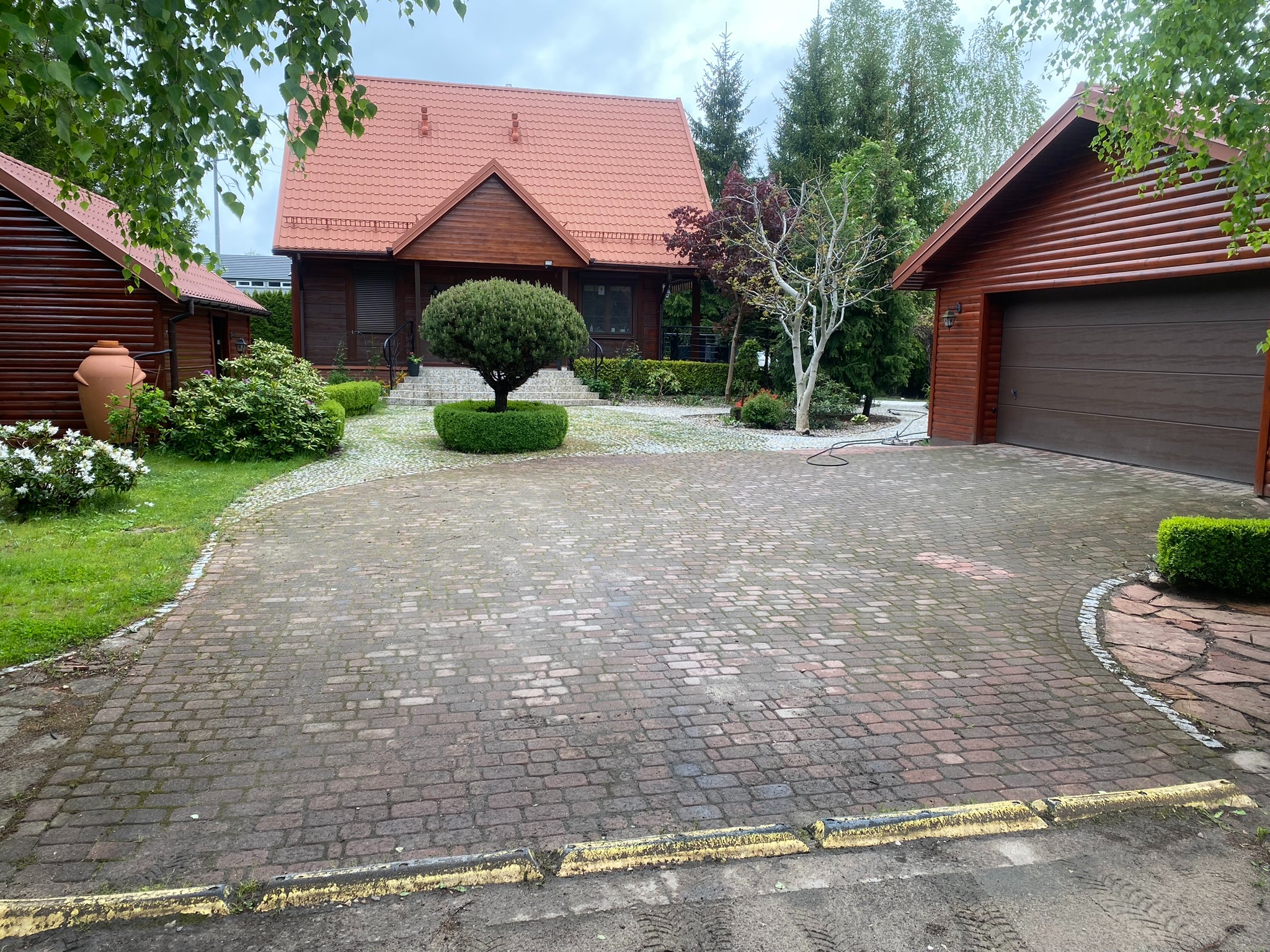 Rozległy podjazd z kostki brukowej prowadzi do drewnianego domu z czerwonym dachem, otoczonego zielenią. Widoczny garaż i elementy dekoracyjne ogrodu.