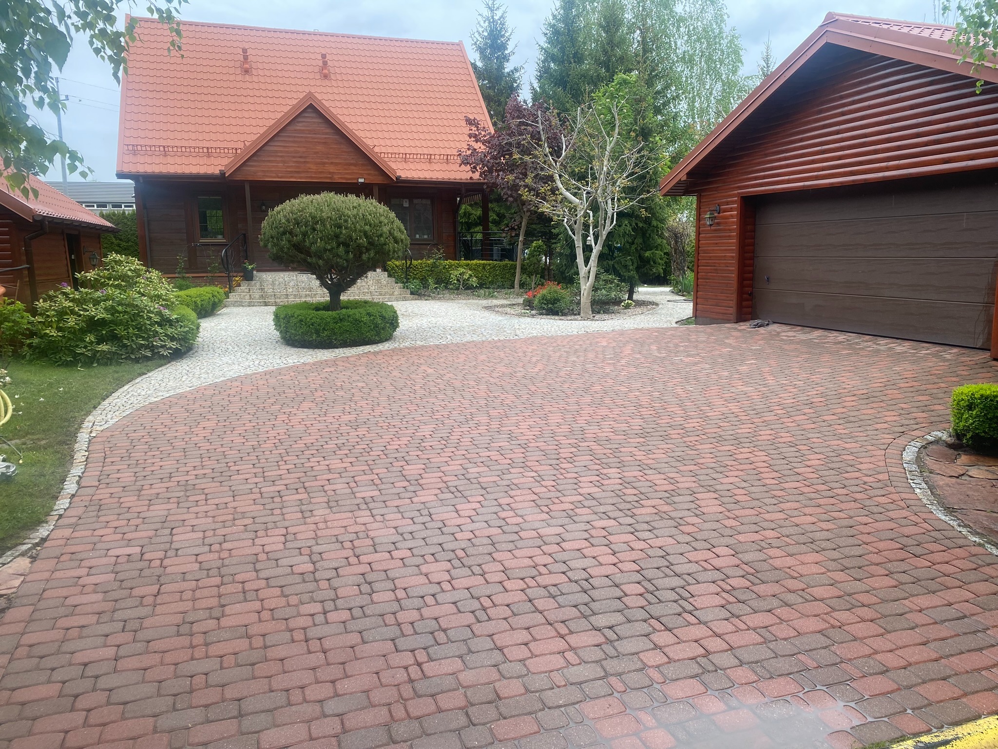 Ceglana kostka brukowa na podjeździe prowadzącym do drewnianego domu z czerwonym dachem i garażu. Uwagę przykuwa okrągły krzew i zadbana zieleń.