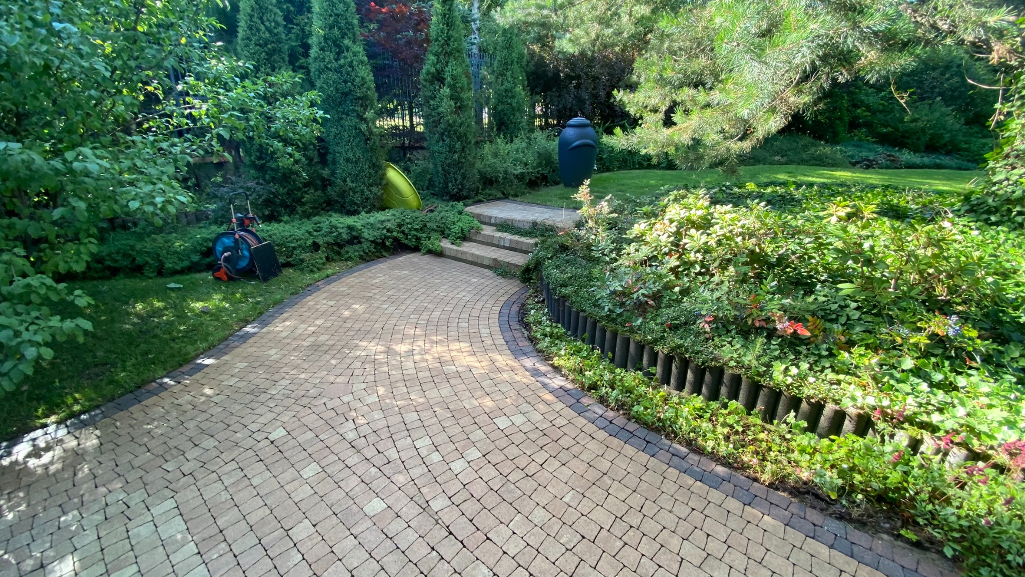 Ułożona kostka brukowa tworząca ścieżkę w ogrodzie, z widocznymi schodami i elementami dekoracyjnymi. W tle bujna zieleń i narzędzie ogrodnicze.