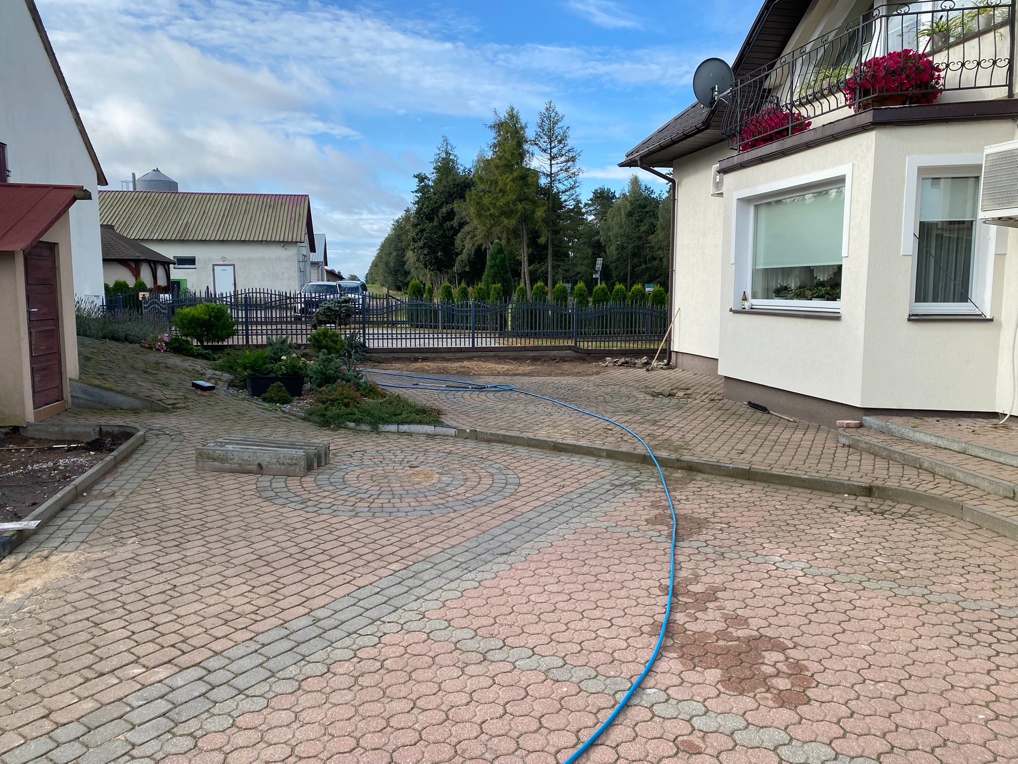 Częściowo umyta kostka brukowa na podjeździe domu jednorodzinnego, z widocznym wężem ogrodowym. Różnica w kolorze kostki wskazuje na postęp prac.