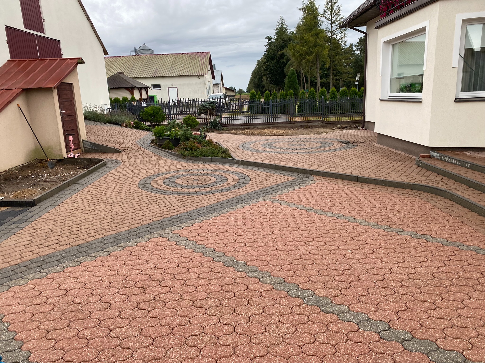 Ułożona kostka brukowa w odcieniach czerwieni i szarości tworzy wzór na podjeździe. Widoczne budynki w tle i elementy zieleni.