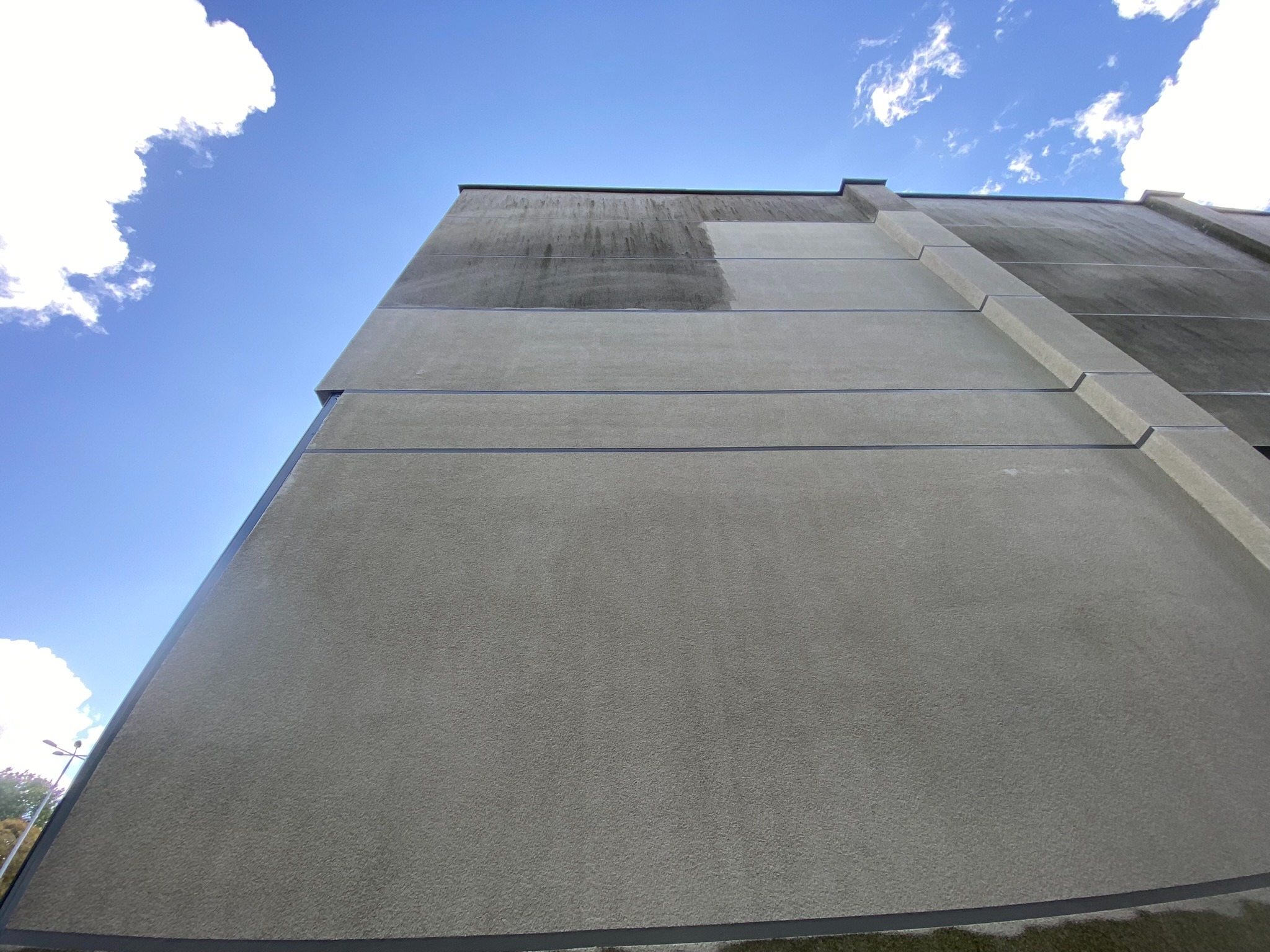 Częściowo umyta, szara elewacja budynku z widoczną różnicą w czystości, na tle błękitnego nieba z chmurami. Ujęcie z dołu podkreśla pionowość ściany.