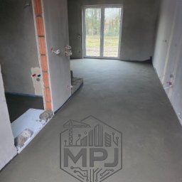 MPJ Martyna Jarota Posadzki betonowe Wylewki styrobetonowe - Świeżo wylana, gładka posadzka jastrychowa w surowym wnętrzu z widocznymi instalacjami elektrycznymi i oknem na ogród. Na posadzce szary znak wodny firmy MPJ.