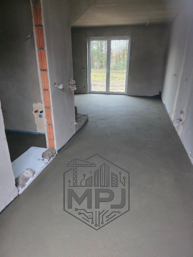 Świeżo wylana, gładka posadzka jastrychowa w surowym wnętrzu z widocznymi instalacjami elektrycznymi i oknem na ogród. Na posadzce szary znak wodny firmy MPJ.