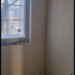 SPEED - PUB RADOSŁAW BUCHALIK - Wnętrze po tynkowaniu maszynowym: gładkie, beżowe ściany, okno z folią ochronną, widoczne listwy przypodłogowe. Surowy stan wykończenia, przygotowany do dalszych prac.