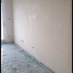 SPEED - PUB RADOSŁAW BUCHALIK - Gładka, beżowa ściana po tynkowaniu maszynowym, widoczne otwory na gniazdka, surowy beton na podłodze, listwa przypodłogowa. Świeżo wykończone wnętrze.