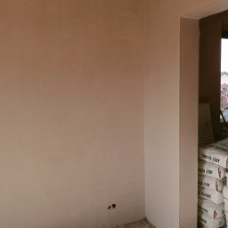 SPEED - PUB RADOSŁAW BUCHALIK - Świeżo otynkowana ściana w rogu pokoju, z widocznymi workami gipsu Dolina Nidy przy oknie. Widoczne niedoskonałości w dolnej części ściany.