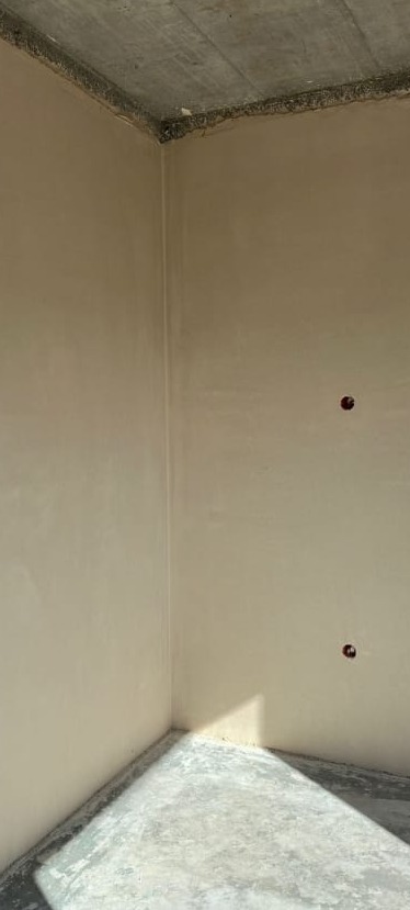 Wnętrze po tynkowaniu maszynowym: gładkie, beżowe ściany, surowy betonowy sufit i podłoga. Widoczne dwa otwory na gniazdka elektryczne.