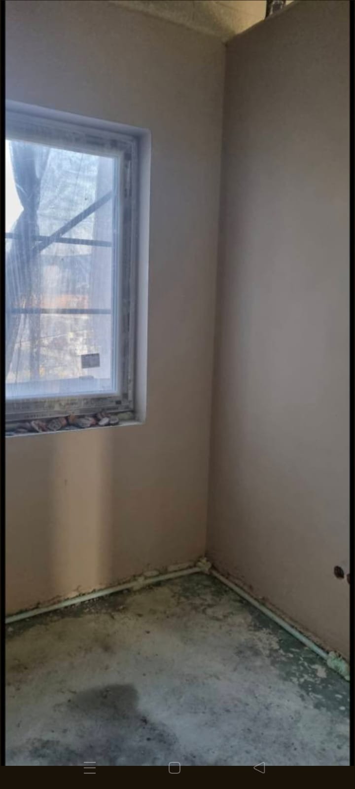 Wnętrze po tynkowaniu maszynowym: gładkie, beżowe ściany, okno z folią ochronną, widoczne listwy przypodłogowe. Surowy stan wykończenia, przygotowany do dalszych prac.