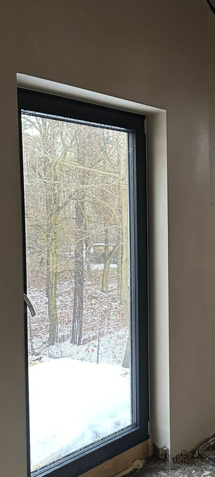 Świeżo otynkowana ściana wewnątrz budynku z widokiem na zimowy krajobraz przez okno. Szare okno, gładka powierzchnia tynku. Widoczne ślady po pracach wykończeniowych.