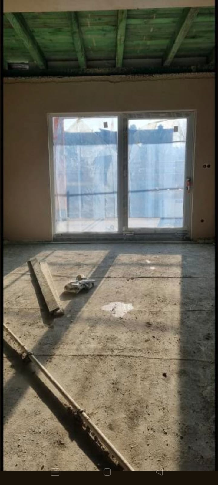 Wnętrze w trakcie prac wykończeniowych: ściany po tynkowaniu, okno zabezpieczone folią, widoczne narzędzia i surowa podłoga. Drewniana konstrukcja stropu.