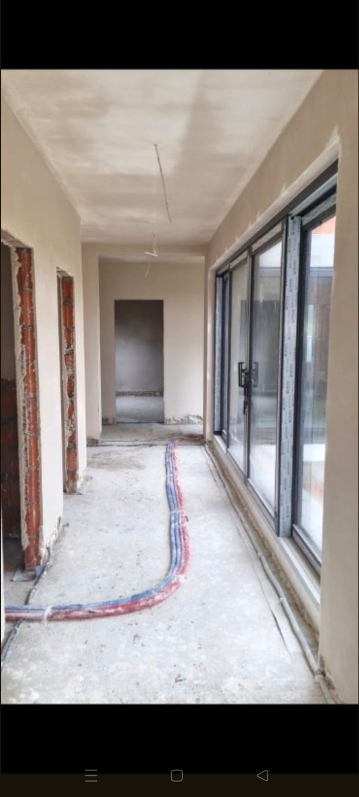 Wnętrze w stanie deweloperskim po tynkowaniu: gładkie ściany, okno przesuwne, instalacja podłogowa. Widok na surowy korytarz z otworami drzwiowymi.