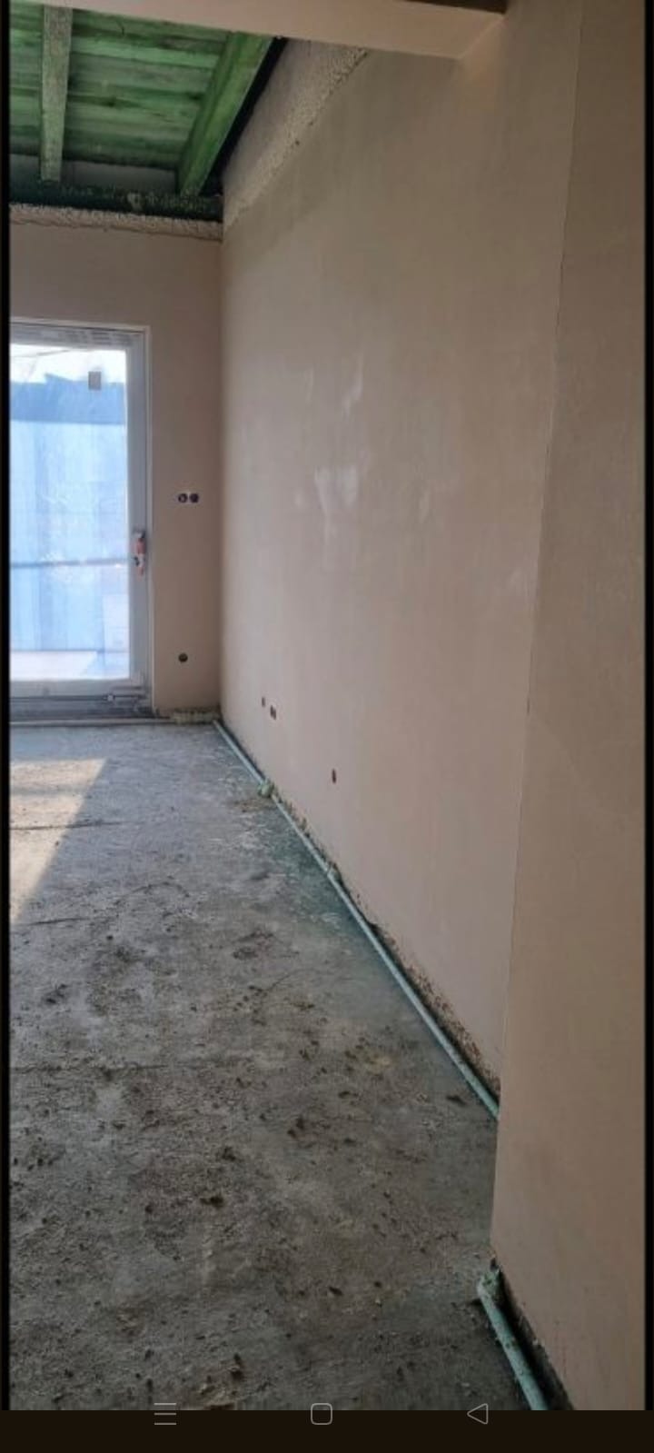 Wnętrze po tynkowaniu maszynowym: gładkie, jasne ściany, surowy beton na podłodze, widoczne instalacje przy podłodze, okno z folią ochronną.