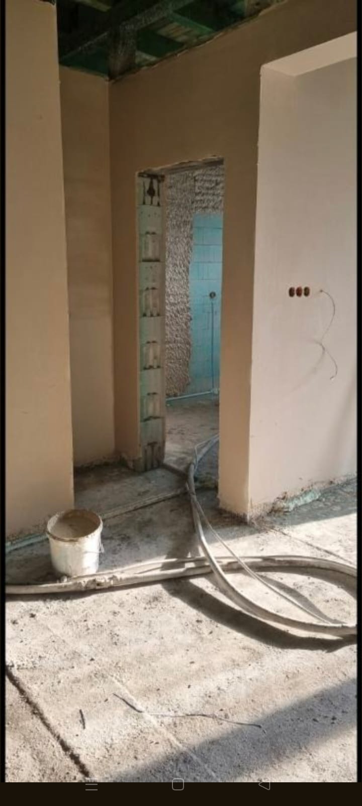 Wnętrze w trakcie prac wykończeniowych: ściany pokryte świeżym tynkiem, widoczne przewody i wiadro z zaprawą na podłodze. W tle surowe ściany i instalacje.