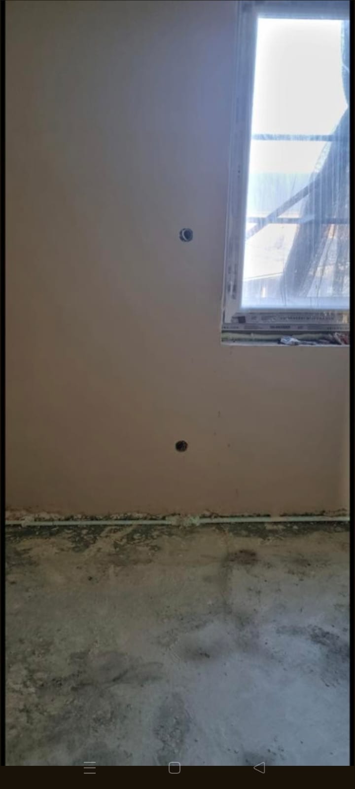 Ściana po nałożeniu tynku maszynowego, widoczne dwa otwory na instalacje. Przy oknie zabezpieczenia folią malarską. Podłoga surowa, przygotowana do dalszych prac.