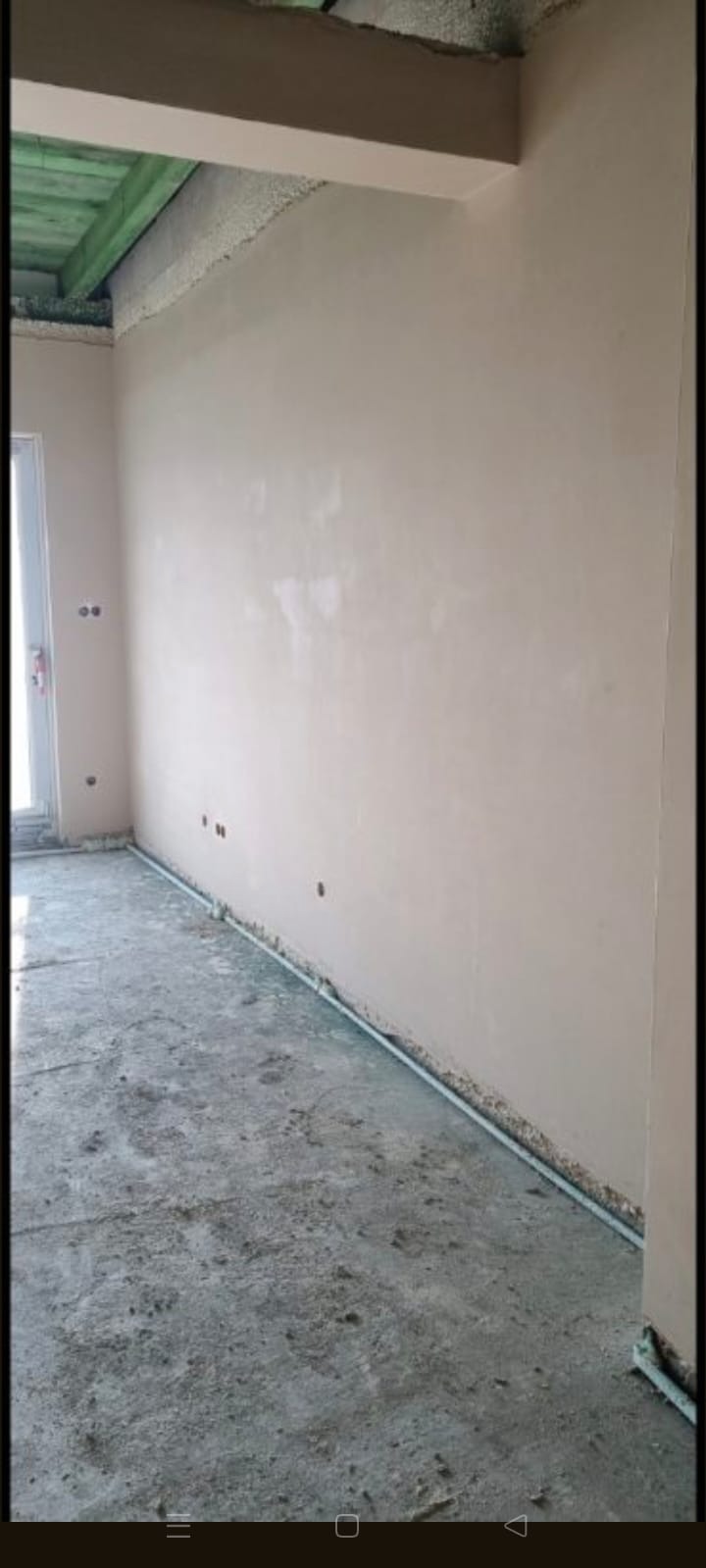 Gładka, beżowa ściana po tynkowaniu maszynowym, widoczne otwory na gniazdka, surowy beton na podłodze, listwa przypodłogowa. Świeżo wykończone wnętrze.