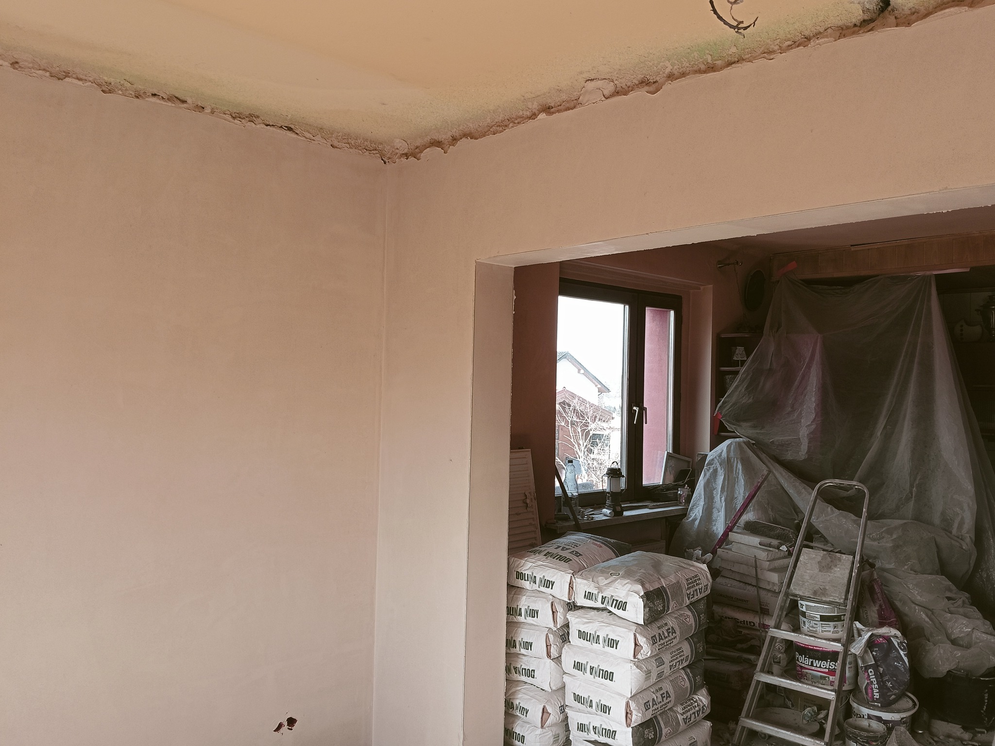 Wnętrze w trakcie prac tynkarskich: gładka ściana po tynkowaniu, worki z zaprawą Dolina Nidy, drabina i okno w tle. Widoczne niedoskonałości na suficie.