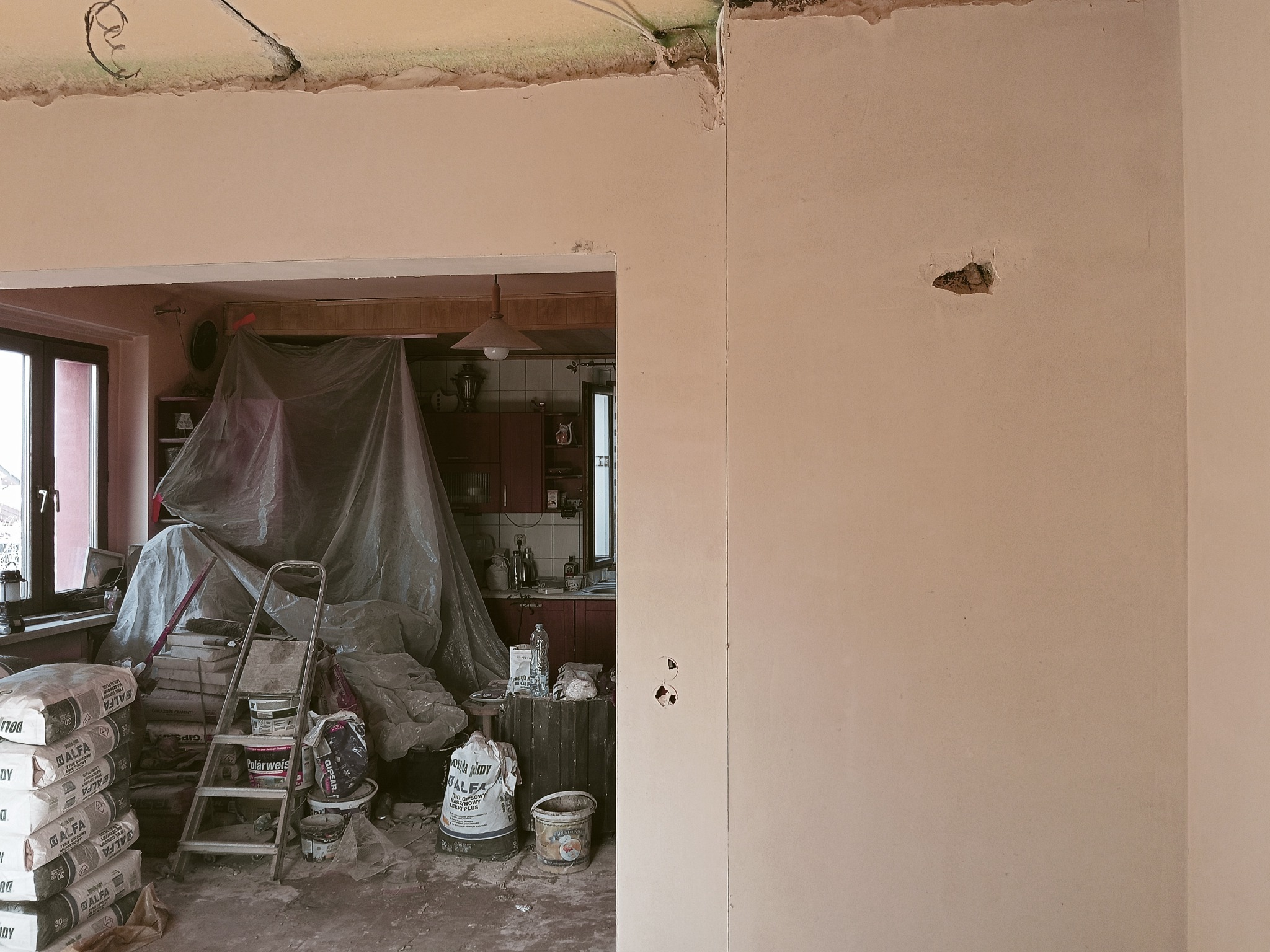 Wnętrze w trakcie prac tynkarskich: worki z zaprawą, drabina, zakryta folią kuchnia w tle, świeżo otynkowana ściana z widocznymi niedoskonałościami.