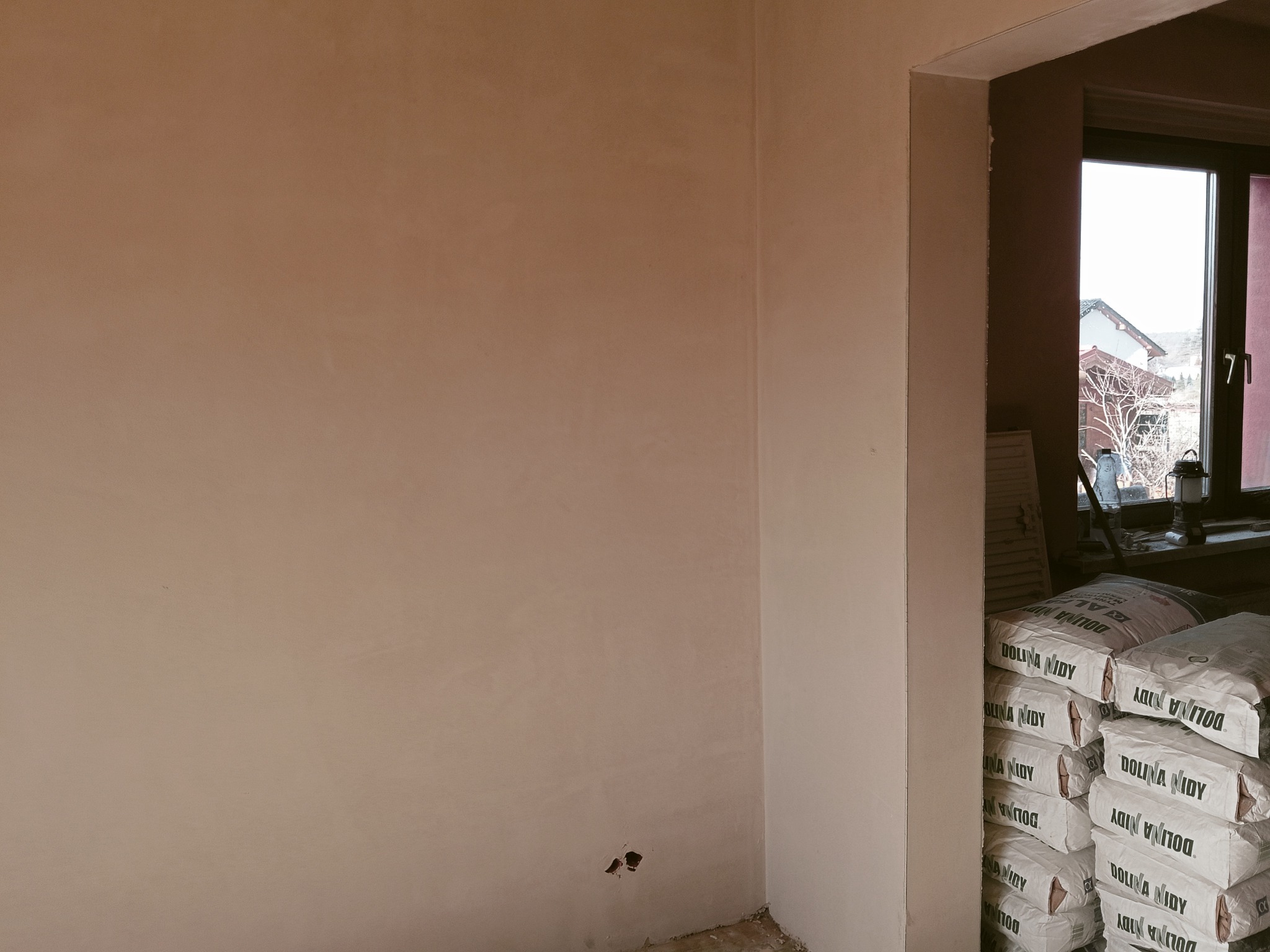 Świeżo otynkowana ściana w rogu pokoju, z widocznymi workami gipsu Dolina Nidy przy oknie. Widoczne niedoskonałości w dolnej części ściany.