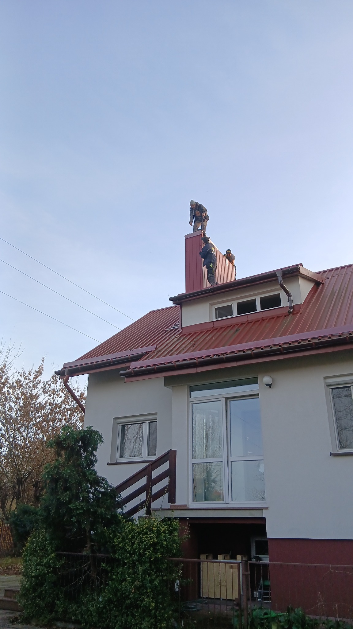 Trzech dekarzy na dachu domu z czerwoną blachodachówką, pracujących przy kominie. Widoczne rynny i fragment elewacji budynku. Prace na wysokości.