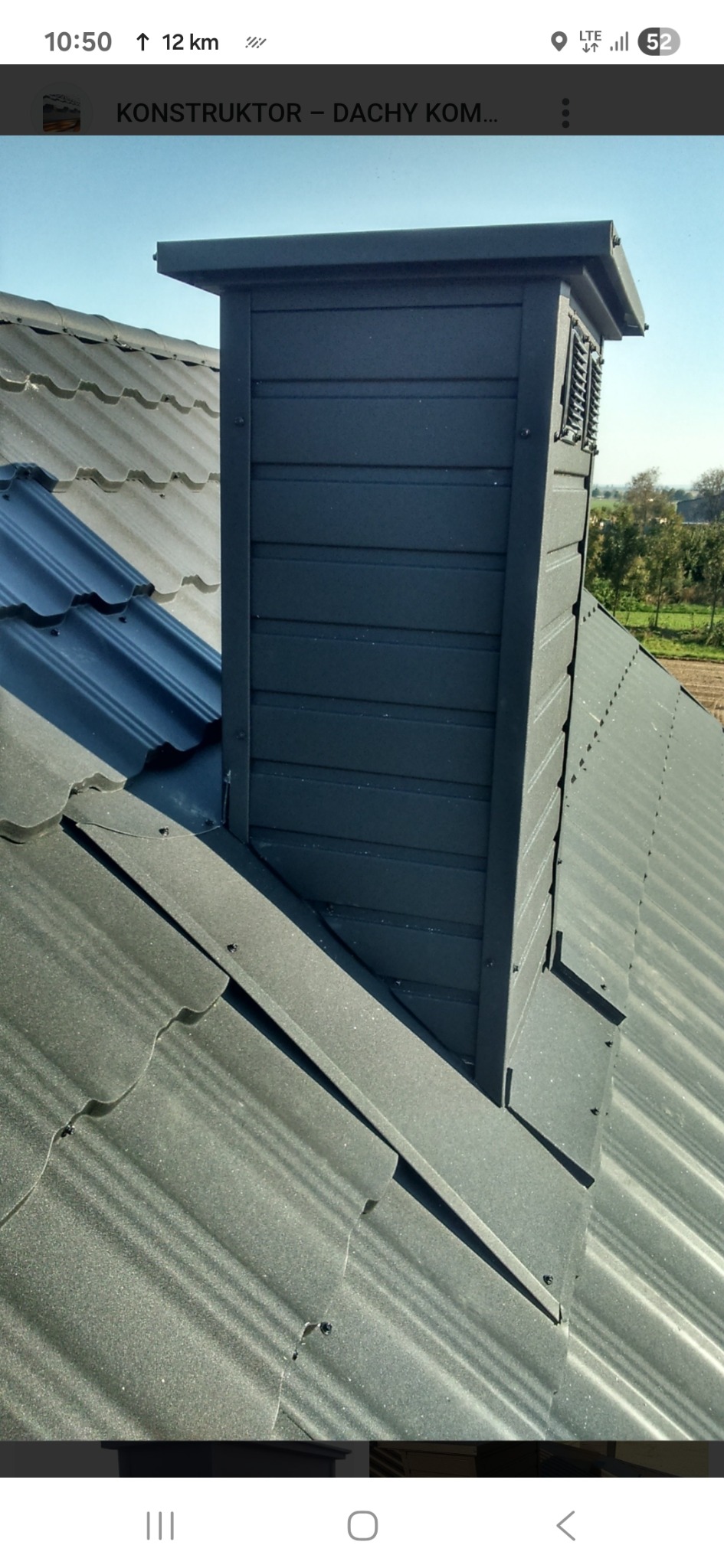 Szary komin wentylacyjny z blachy na dachu z blachodachówki w odcieniach szarości, widok z góry, na tle zieleni drzew i pola.