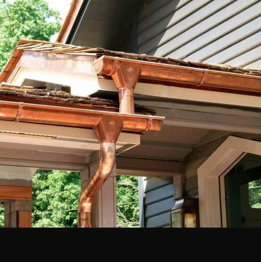 Miedziane rynny z dekoracyjnymi złączkami na tle szarej elewacji domu z widokiem na zieleń. Detal architektoniczny, eleganckie wykończenie dachu.