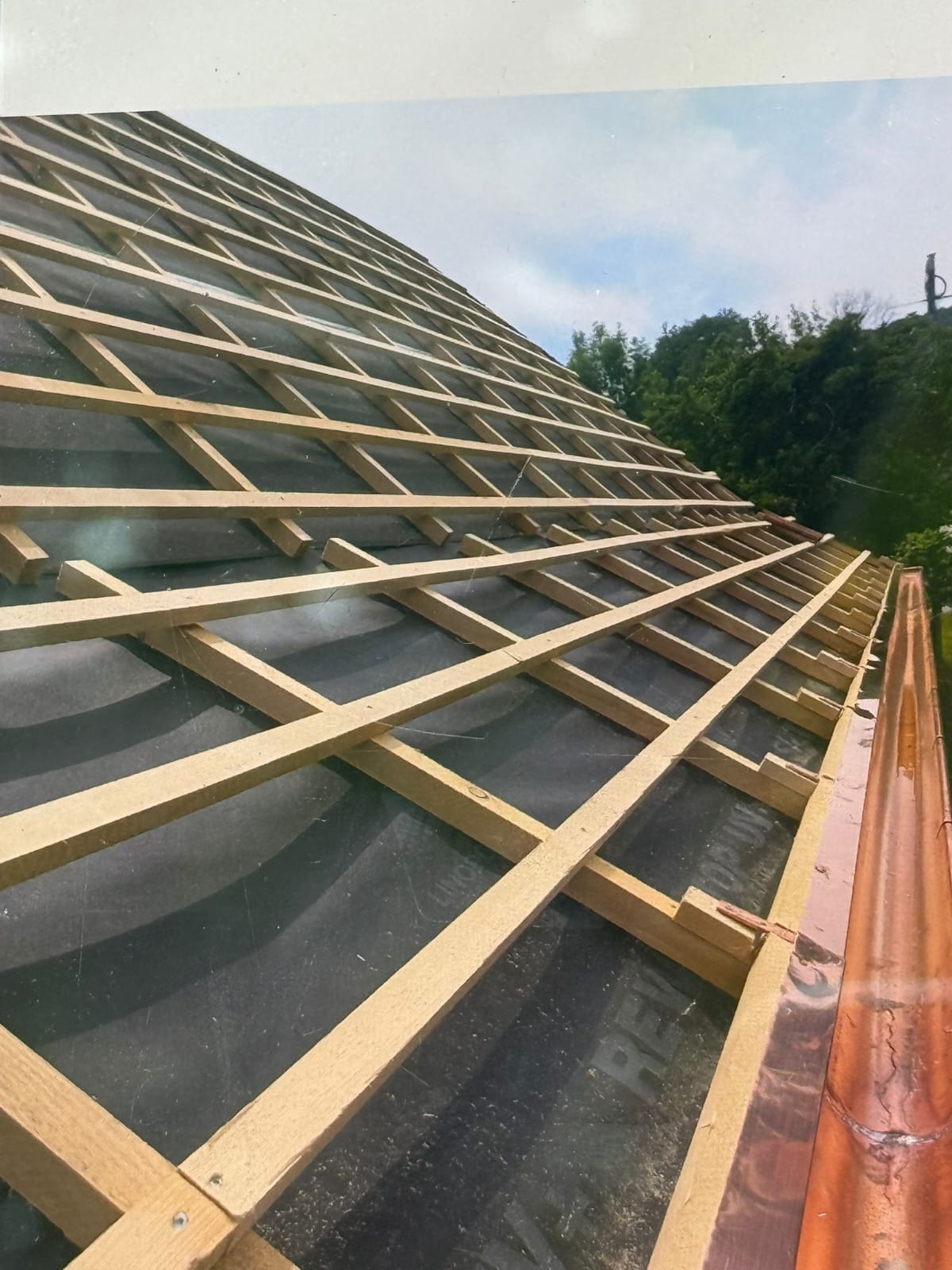 W trakcie budowy dachu: drewniana więźba dachowa na czarnej membranie, widoczna miedziana rynna. Ujęcie z dołu, w tle zieleń drzew i fragment nieba.