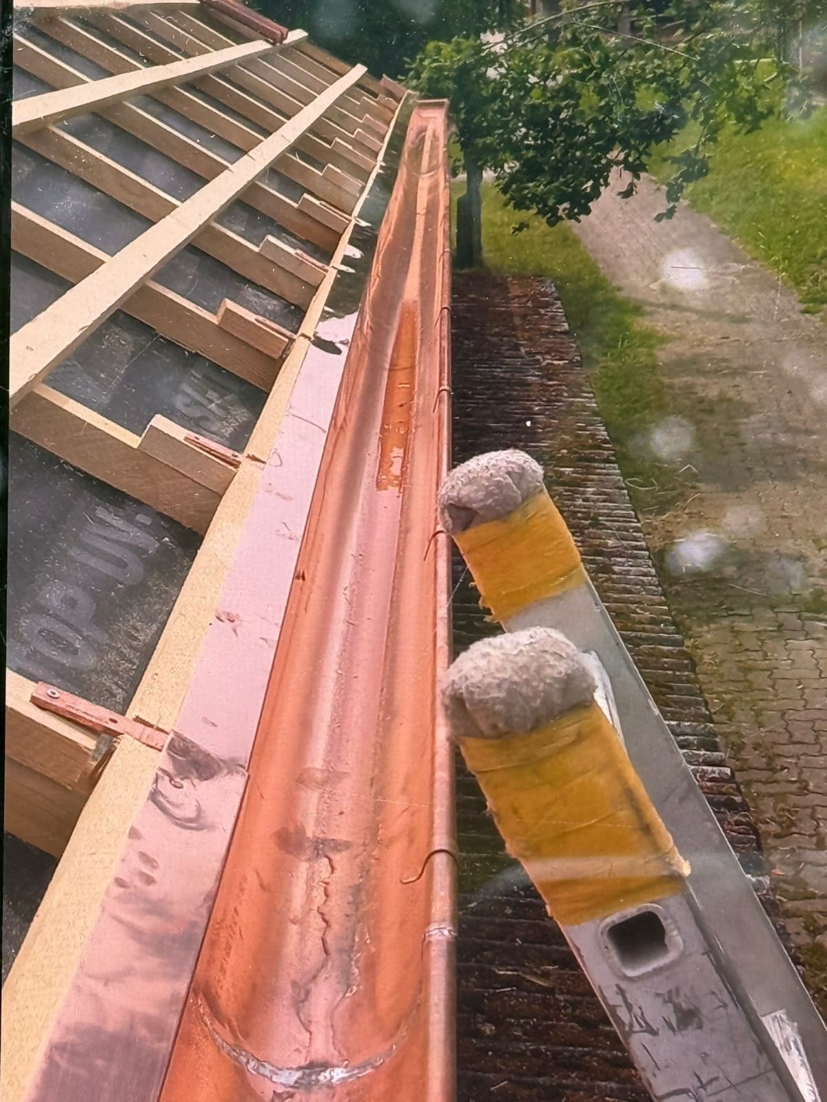 Miedziana rynna zamontowana na dachu z widoczną więźbą, z boku oparta drabina z zabezpieczeniem. Widok z góry na dach i otoczenie.