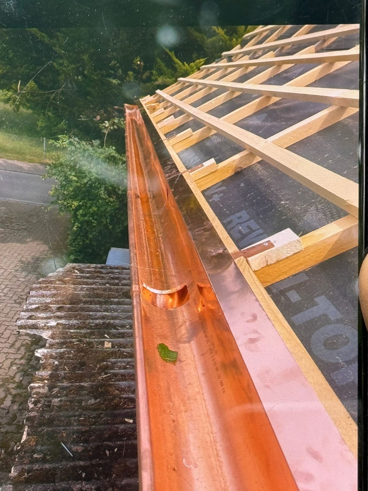 Miedziana rynna zamontowana na dachu z widoczną więźbą dachową i fragmentem starego pokrycia. Na dnie rynny leży zielony listek. Ujęcie z góry.