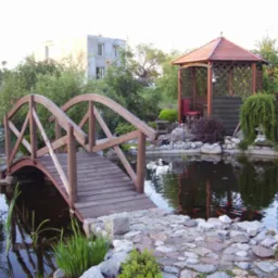 Oczko wodne z drewnianym mostkiem i altaną w ogrodzie. Woda odbija niebo i architekturę, dodając głębi. Kamienne brzegi i roślinność tworzą naturalny klimat.