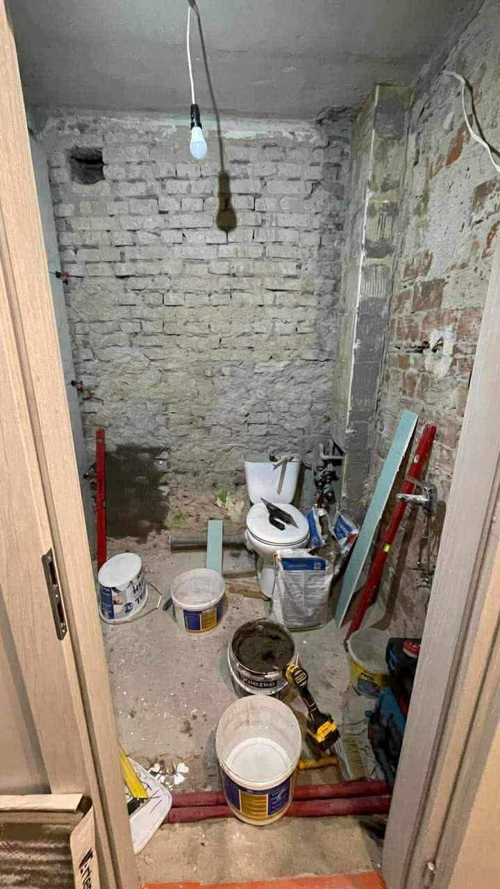 Łazienka w trakcie remontu: odsłonięta cegła, wisząca żarówka, narzędzia i farby. Widok przez otwarte drzwi, ukazujący etap prac przed wykończeniem.