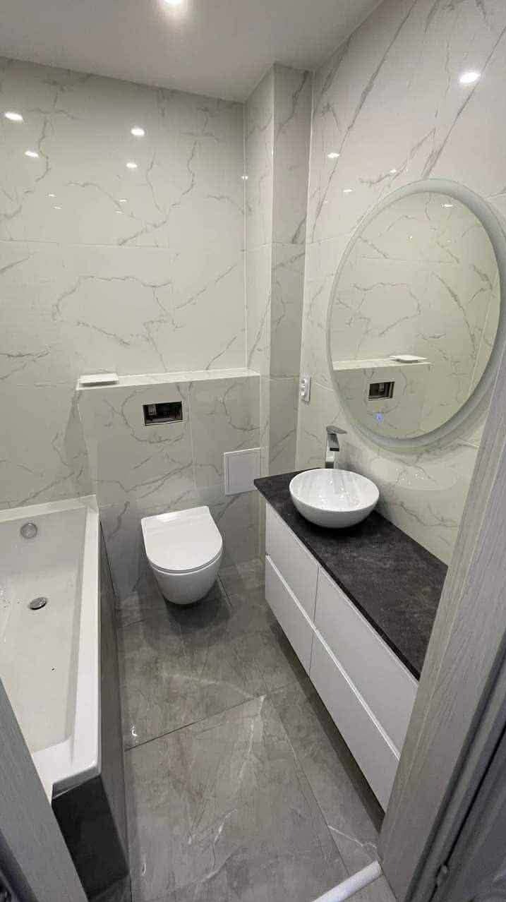 Nowoczesna łazienka po remoncie: marmurowe płytki, okrągłe lustro z podświetleniem, biała umywalka nablatowa i podwieszana toaleta. Minimalistyczny design.